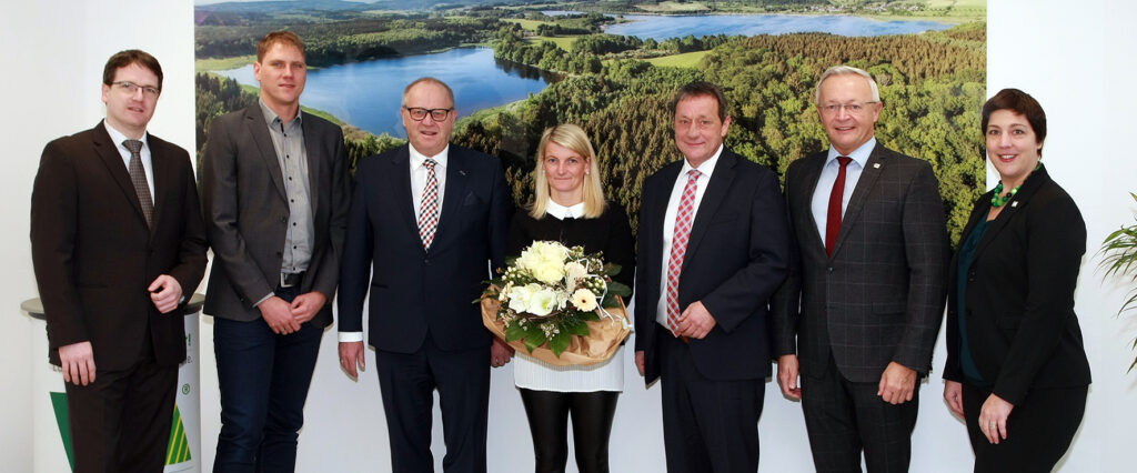 Man erkennt fünf Männer und zwei Frauen, die alle elegant gekleidet vor einem großen Bild stehen. Das Bild zeigt einen See mit viel grüner Natur. Eine Frau steht in der Mitte und hält einen Blumenstrauß.