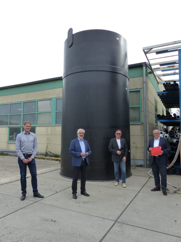 vier Personen in eleganter Kleidung, die draußen vor einem großen, schwarzen Zylinder stehen, der vor einer Industriehalle befestigt ist.