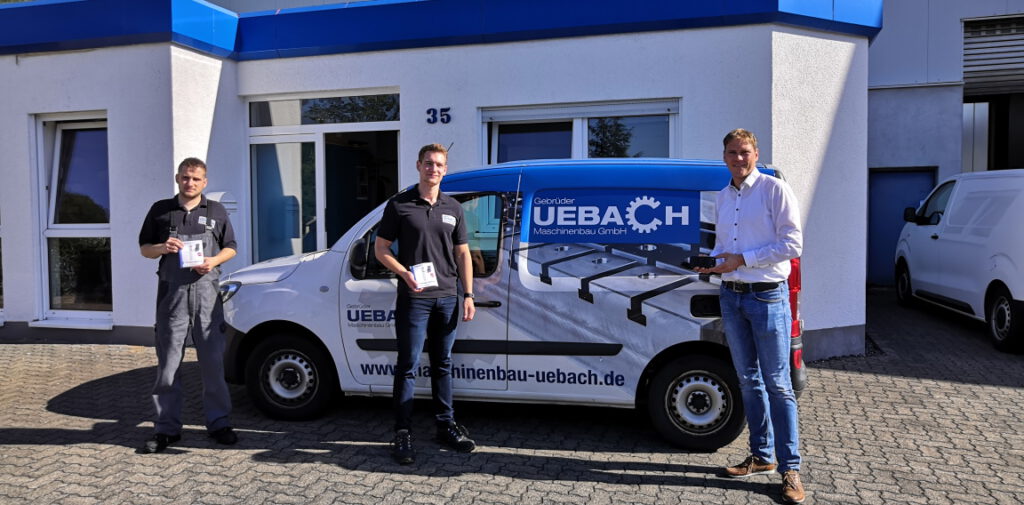 Auf dem Bild stehen die zwei Geschäftsführer der Firma Uebach und der Leiter der Wirtschaftsförderung vor einem Firmenwagen der Firma Uebach. Das Auto steht vor dem weißen Firmengebäude mit blauem Dach.